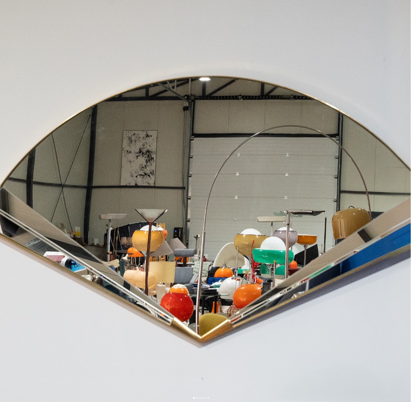 A brass fan-shaped mirror on a white wall, reflecting the prop hall and its rows of coloured lamps