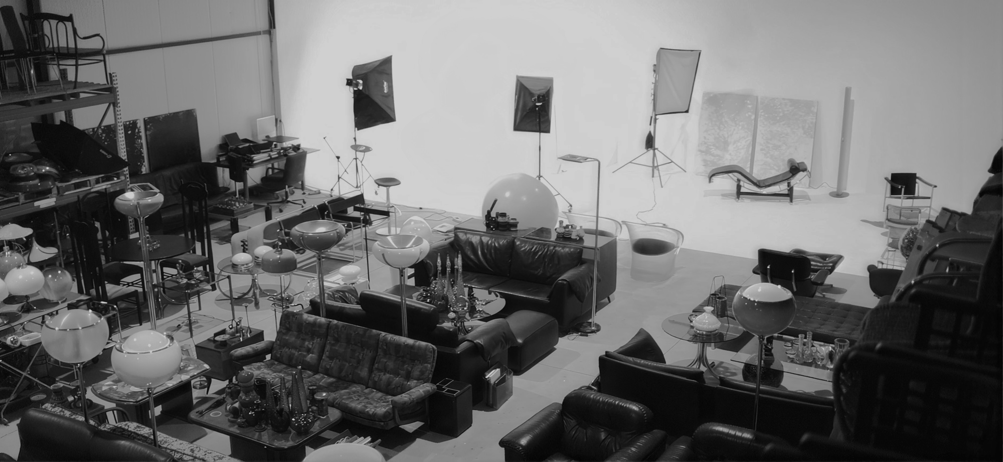 The studio floor seen end to end — leather sofas, globe lamps and glass tables massed in front of the lit white cyc, softboxes rigged along the wall.